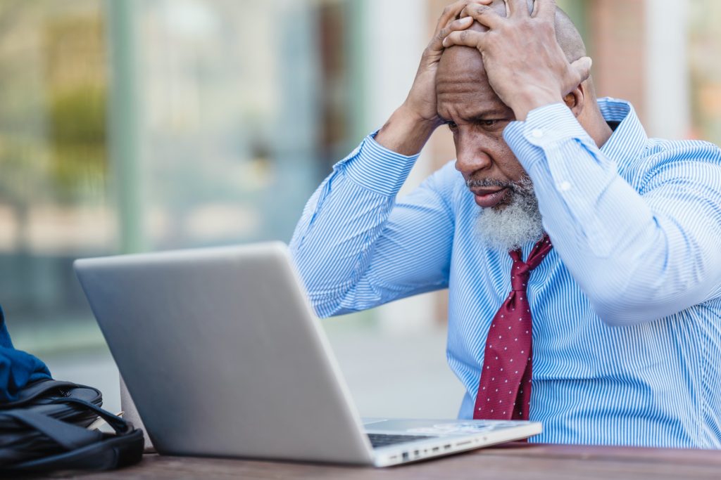 An older man with his hands on his head, looking at his laptop.