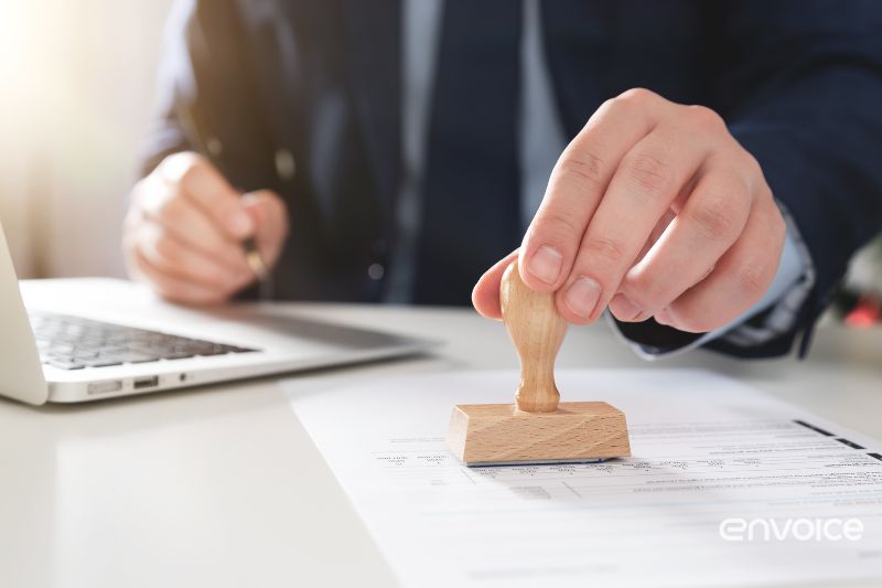 A person in a business suit stamps a document on a desk with a laptop nearby, symbolizing the approval process in invoice handling