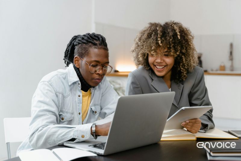 Two colleagues work together in an office, one using a laptop and the other holding a tablet, with smiles indicating collaboration and efficiency