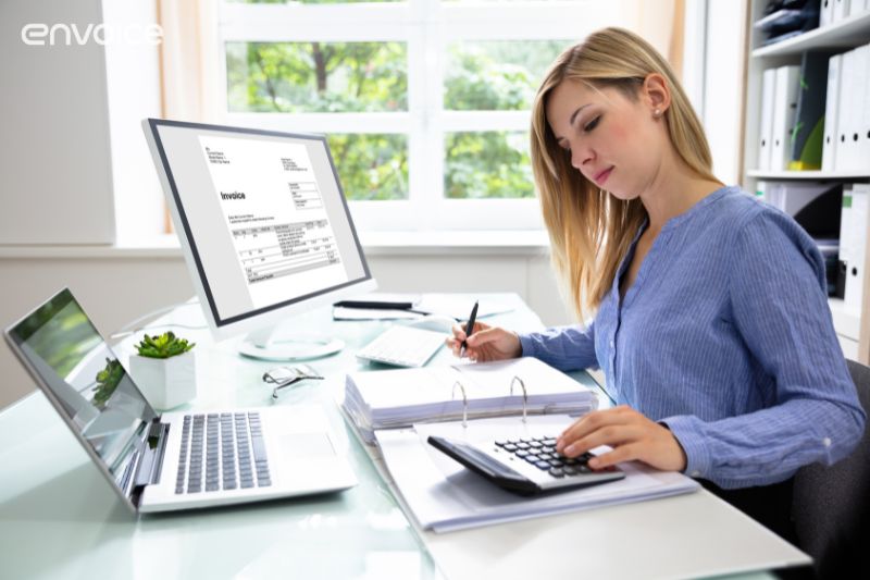A focused woman works at a desk reviewing documents in a binder, with an invoice displayed on a computer monitor exploring alternative accounting software options.