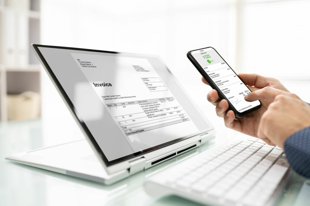 A person sitting at a desk is using a laptop and holding a smartphone displaying the same invoice representing the convenience of accessing and managing invoices on multiple devices