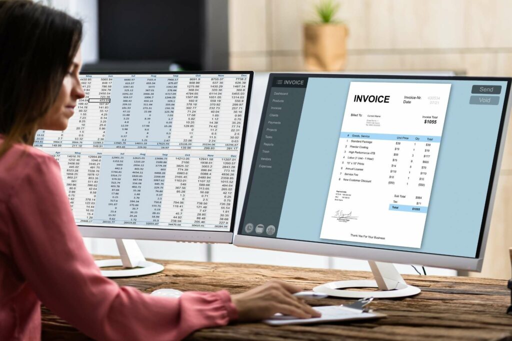 A woman sits at a desk working on two computer monitors displaying an invoice and a detailed expense spreadsheet