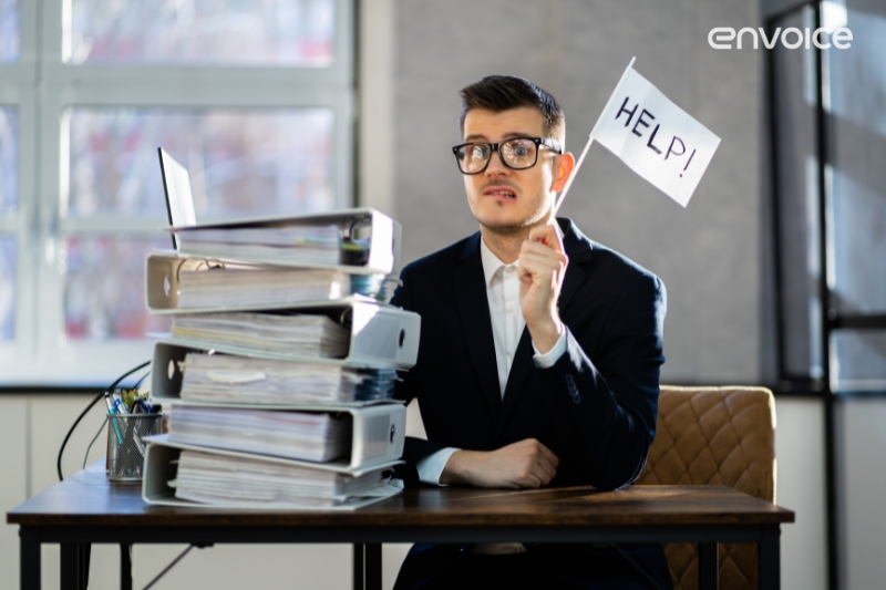 Overwhelmed businessman sitting at a desk with a large stack of binders, holding a small flag that says 'HELP!' - representing the need for streamlined payment processes.