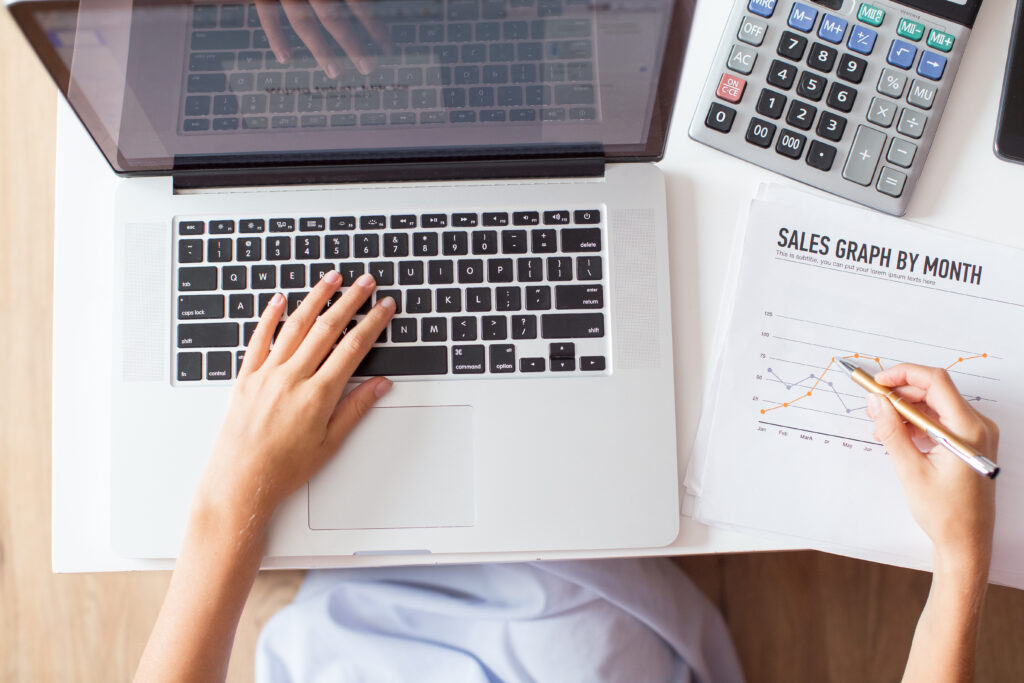 A person working on a laptop, analyzing a sales graph on a printed document.